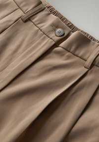 Beige, gefaltete Hose mit elastischem Bund, ausgestattet mit einem Knopfverschluss und einem glatten, strukturierten Stoff.