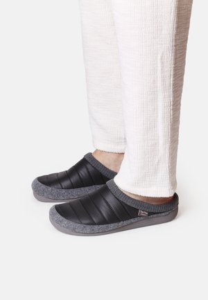 Zapatillas de casa deslizables en negro y gris con un diseño acolchado, material suave similar a fieltro y suela de goma. Acompañadas de pantalones blancos texturizados.