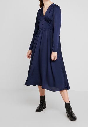 Navy blue dress with long sleeves, V-neck, pleated detailing, and buttons down the front. Accompanied by black ankle boots.
