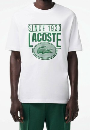 Biały bawełniany T-shirt z zielonym napisem "LACOSTE" i logo, okrągły dekolt, krótkie rękawy, z graficznymi detalami z roku 1933.