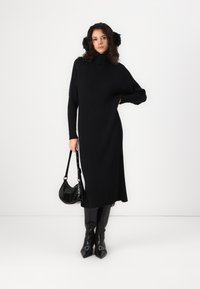 Black ribbed knit dress with a high collar, side slit, and long sleeves. Paired with knee-high black boots and a small black handbag.