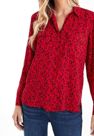 Femme portant un chemisier rouge à manches longues avec un imprimé léopard noir et un jean bleu, debout devant un fond blanc.