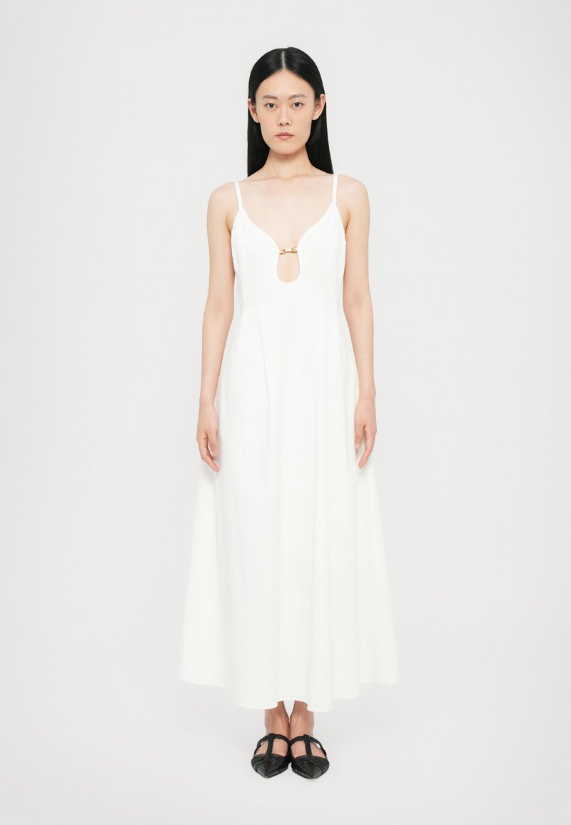 Vestido maxi blanco con un profundo escote en V y un acento dorado en el pecho. Cuenta con tirantes finos y un diseño suave y fluido. Zapatos planos negros.