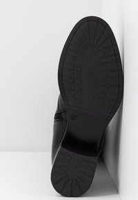 Musta kenkänsija, jossa on syvät kuvioinnin ja koko 37 merkintä, kuvattu valkoista kulmaa vasten.