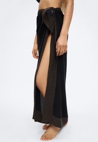 Γυναίκα που φοράει μαύρη φούστα με άνοιγμα στο πλάι και διακοσμητικό μοτίβο στο τελείωμα, όρθια ξυπόλυτη μπροστά σε απλό φόντο.