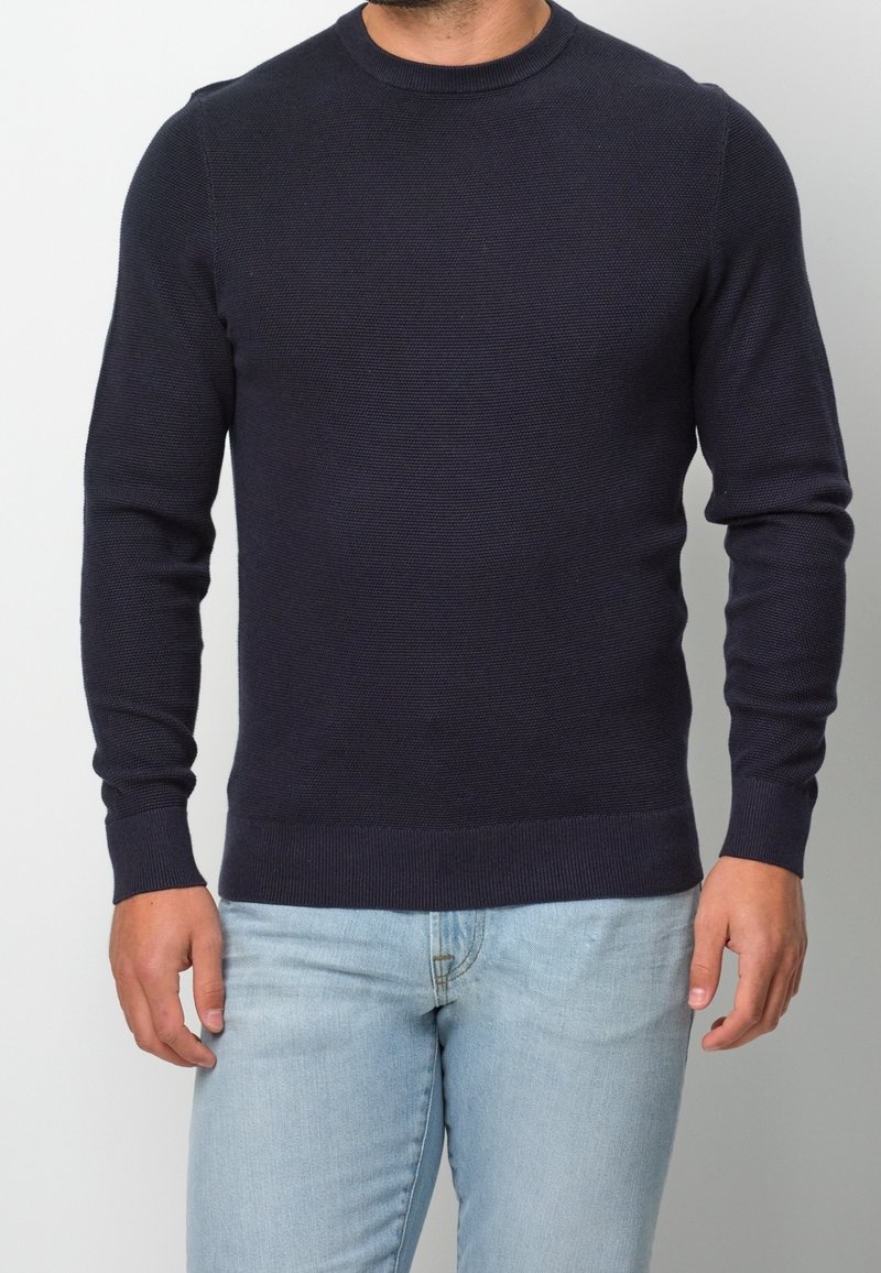 Homme portant un pull en maille texturée bleu marine foncé et un jean bleu clair, debout devant un fond clair uni.