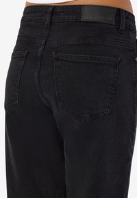 Črne denim hlače z zadnjimi žepi in pasnimi zankami, oblečene na osebi, ki prikazuje območje pasu in zgornjega dela stegna.