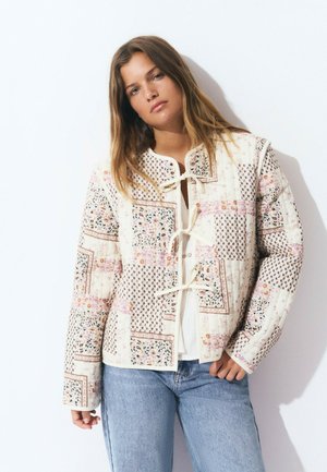 Chaqueta acolchada en crema con patrones florales y geométricos, presenta un escote redondo y un cierre frontal atado. Tela ligera con textura.