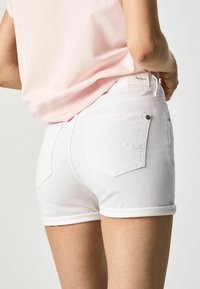 Pantalones cortos de denim blanco con dobladillos enrollados, que cuentan con bolsillos traseros y una pequeña etiqueta de la marca en la cinturilla. Textura suave y estructurada.