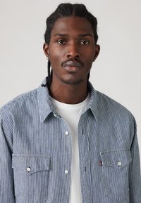 Ριγέ πουκάμισο σε μπλε και λευκό με κουμπιά, δύο τσέπες στο στήθος και κλασικό γιακά, φορεμένο πάνω από λευκό μπλουζάκι. Ομαλή υφή υφάσματος.