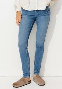 Jeans skinny en denim bleu clair, présentant une coupe classique. Associés à des sabots en suède marron accentués par des clous en métal.
