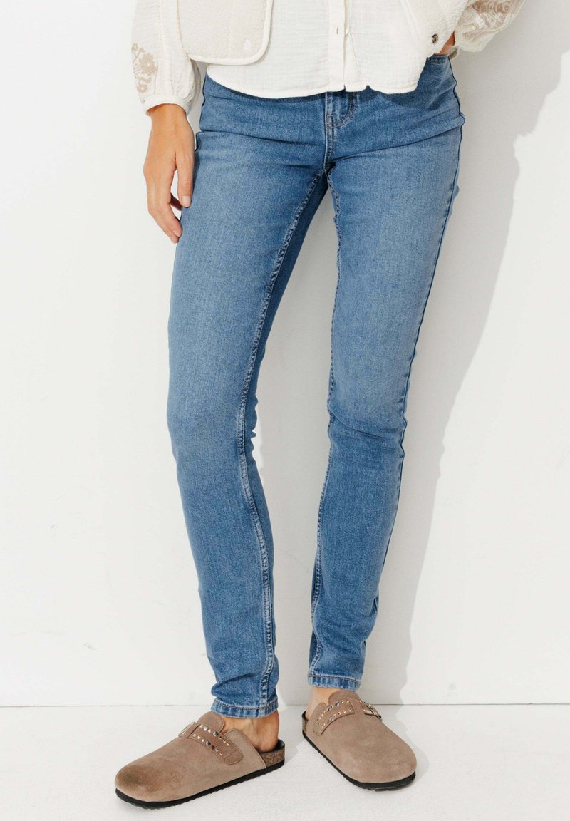 Jeans skinny en denim bleu clair, présentant une coupe classique. Associés à des sabots en suède marron accentués par des clous en métal.
