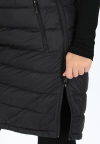 Svart pufferväst med quiltat mönster; har en sidodragkedjeficka; slät textur; hand som greppar dragkedjan.