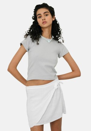 Ung kvinde med krøllet hår iført en lys grå tætsiddende crop top og hvid omslagsnederdel, poserer med hænderne på hofterne foran en ensfarvet baggrund.