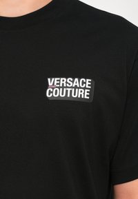 Juodos medvilnės marškinėliai su akcentu baltu ir rožiniu "VERSACE JEANS COUTURE" logotipu kairėje krūtinės pusėje. Trumpomis rankovėmis, apvalia iškirpte.
