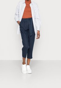 Pantalones cortos de marinero con cintura atada, combinados con una camiseta de color óxido y una camisa de botones a rayas blancas, usados con zapatillas blancas.