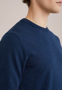 Tamsiai mėlynas apvalios iškirptės megztinis, pagamintas iš minkštos, tekstūrinės medžiagos, su prigludusia forma ir subtiliais raukšlių akcentais ant iškirptės ir pečių.