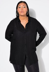 Frau in einem schwarzen, gestreiften, geknöpften Hemd, großen Creolen und schwarzen Leggings mit kleinen weißen Punkten vor einem weißen Hintergrund.