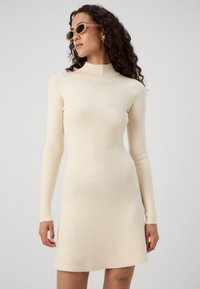 Vestito lavorato a maglia color crema con colletto alto, maniche lunghe e una silhouette aderente che si allarga all'orlo. Texture liscia con una leggera lucentezza.