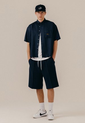 Junger Mann trägt ein marineblaues kurzärmeliges Hemd mit Knöpfen über einem weißen T-Shirt, marineblaue Shorts, weiße Socken, weiße Nike-Sneaker und eine schwarze Kappe.