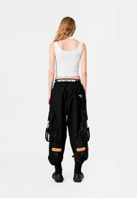 Pantalones cargo negros con múltiples bolsillos y cremalleras, acentos naranjas, cinturilla elástica y una camiseta sin mangas blanca ajustada. Calzado plano y texturizado.