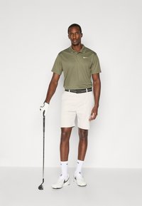 Polo tričko Nike v olivovo zelenej farbe, spárované s béžovými šortkami. Biele tenisky s čiernymi akcentmi. Drží golfovú palicu, na ruke má bielu golfovú rukavicu.