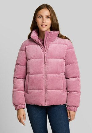 Jeune femme portant une doudoune en velours côtelé rose et un jean bleu foncé, debout devant un fond gris clair uni.