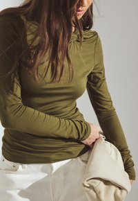 Top a maniche lunghe verde oliva realizzato in tessuto morbido con scollatura arricciata, abbinato a pantaloni color crema, mentre tiene una giacca leggera.