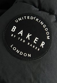 Zwarte stoffen patch met witte reliëftekst met de tekst "BAKER by TED BAKER" en "UNITED KINGDOM LONDON," omlijnd in een cirkelvormig ontwerp.