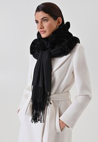Sciarpa nera con colletto in pelliccia sintetica, caratterizzata da dettagli con frange, abbinata a un cappotto bianco. Tessuto morbido e design fluido.