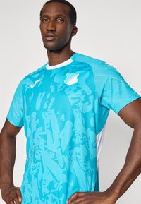 Μπλε αθλητική φανέλα με γεωμετρικό σχέδιο, κοντά μανίκια και λευκές λεπτομέρειες. Φτιαγμένη από ελαφρύ ύφασμα με αναπνεύσιμη υφή.