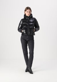 Zwarte pufferjack met een glanzende afwerking, rits aan de voorkant, capuchon en elastische manchetten, gecombineerd met zwarte denim jeans en enkellaarsjes.