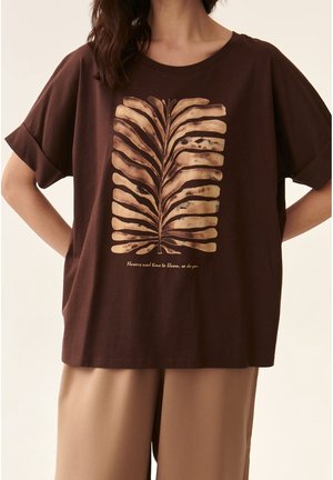 Brun kortærmet skjorte med et beige abstrakt bladmotiv og teksten "Blomster har brug for tid til at blomstre, det har du også," båret med beige bukser.