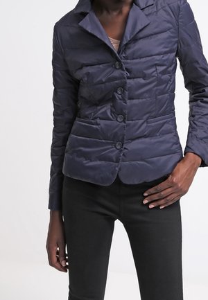 Mujer con una chaqueta acolchada ajustada de color azul marino oscuro con botones y pantalones negros, de pie frente a un fondo claro y liso.