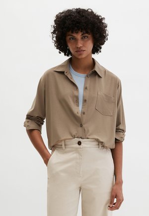 Hellbraunes Hemd mit Knopfleiste und Kragen, einzelne Brusttasche, hochgekrempelte Ärmel, getragen über einem hellblauen, gerippten Oberteil und beiger Hose.