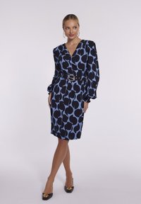 Czarna i niebieska sukienka w wzory z fasonem kopertowym, długimi rękawami, czarnym paskiem i błyszczącymi akcentami. Materiał wydaje się gładki i lekki.