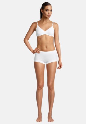 Woman standing barefoot wearing white bra and white boyshort-style underwear against plain light background.