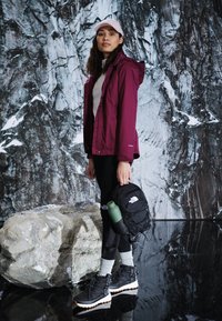 Maroon waterproof jacket over a white top, black leggings, and black hiking boots. Black backpack with green water bottle. Gray rock backdrop.