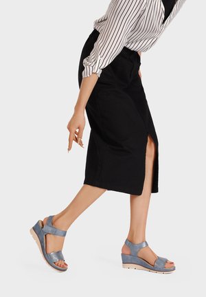 Donna che indossa una gonna a matita nera con spacco laterale e sandali blu con zeppa, solleva una gamba e indica verso il basso con la mano.