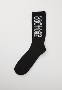 Fekete csős zokni fehér „Versace Jeans Couture” felirattal a bordázott felső részén, sima háttér előtt.