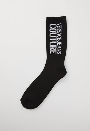 Juodas kojinės su balta „Versace Jeans Couture“ tekstu ant juostos viršaus, priešais vienspalvį foną.