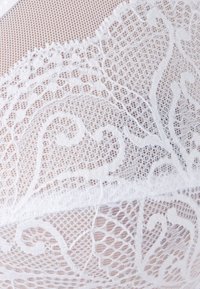 Le tissu en dentelle blanche présente des motifs complexes avec des tourbillons et des détails fins, mettant en valeur un design délicat et texturé sur un fond en maille.