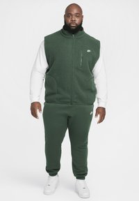 Colete sem mangas em polar verde sobre uma camisa de manga longa branca, combinado com calças de jogging verdes a condizer e t�énis brancos. Apresenta detalhes com o logótipo da Nike.