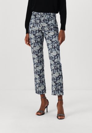 Kobieta ubrana w granatowe krótkie spodnie w kwiatowy wzór, czarną bluzkę z długim rękawem oraz czarne sandały na wysokim obcasie z paskami wokół kostek.