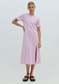 Rosa, kurzärmliges Kleid mit rundem Ausschnitt und seitlichen Schlitzen, aus weichem, glattem Stoff. Kombiniert mit schwarzen, klobigen Sandalen.