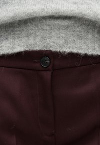 Κοντινή λήψη του τελειώματος ενός πουλόβερ πάνω από τη ζώνη κουμπωμένης στενής μέσης ενός σκούρου βουργουνδί παντελονιού, που δείχνει την υφή της πλέξης και τις λεπτομέρειες του ενδύματος.