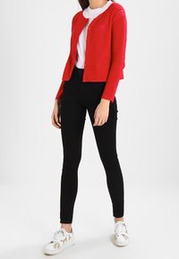 Röd texturerad cardigan med knappar, vit t-shirt, svarta åtsittande byxor och vita sneakers med guldaccenter. Enfärgad bakgrund.