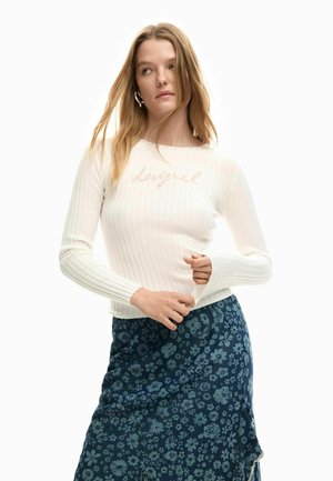 Mladá žena s dlhými blond vlasmi, oblečená v bielom rebrovanom svetri s logom a modrej kvetinovej sukni, pózujúca na bielom pozadí.