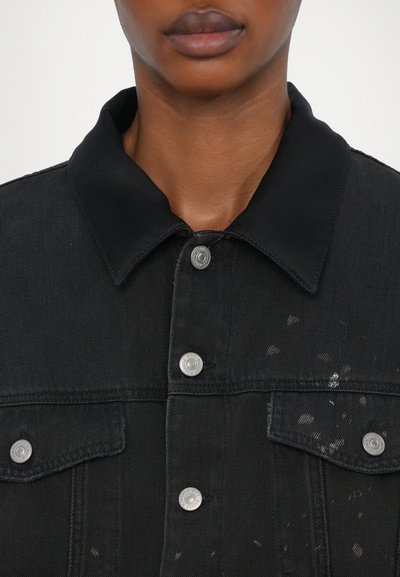 Črna jeans jakna s klasičnim ovratnikom, srebrnimi pritrdilnimi gumbi, sprednjimi žepi in subtilnimi madeži barve na tkanini.
