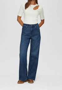 Mujer con una blusa blanca de manga corta y corte en el pecho, jeans anchos de tiro alto en color azul y zapatos de plataforma marrones, de pie con las manos detrás de la espalda.